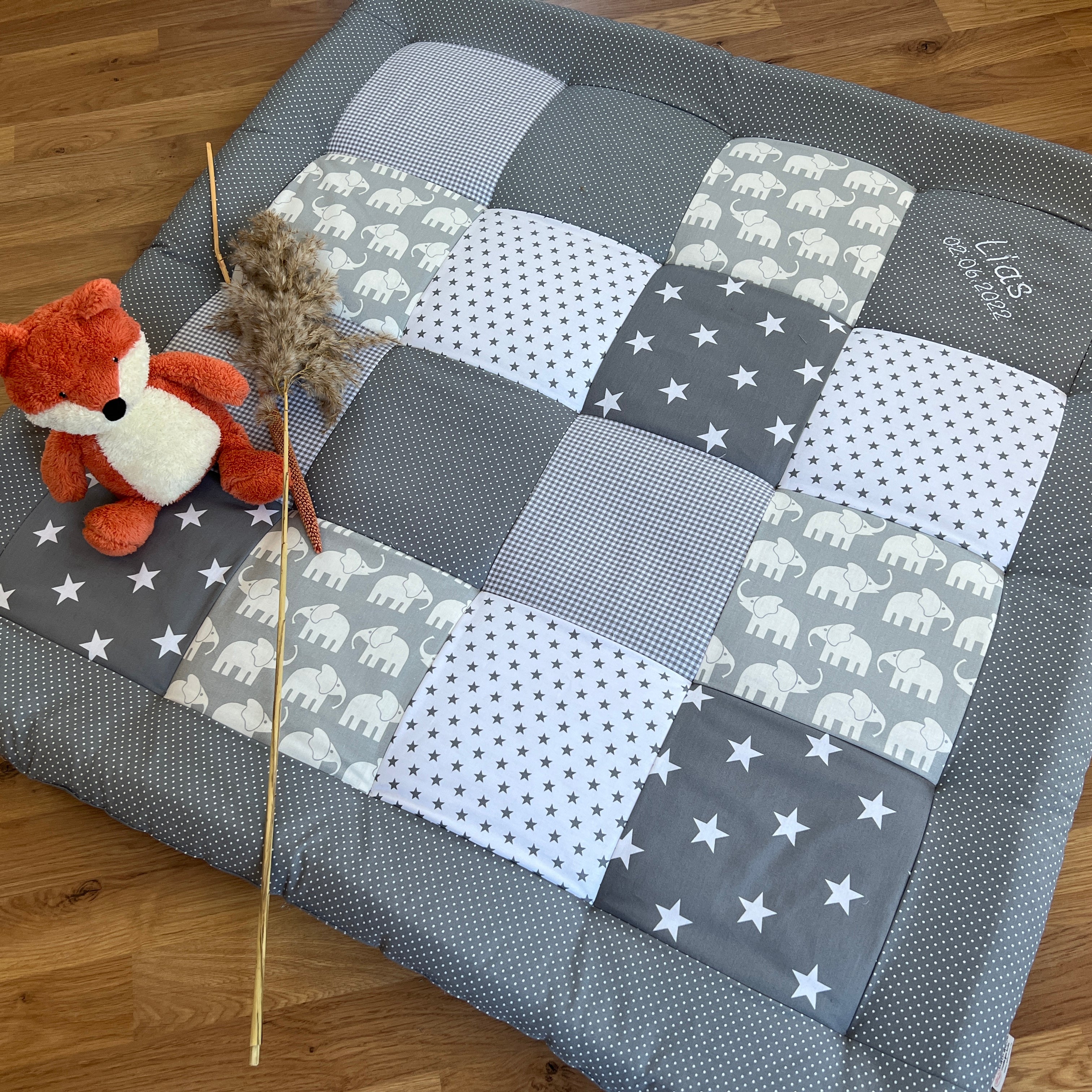 A gray and white patchwork quilt with a stuffed animal fox and a stuffed animal elephant on it, placed on a wooden floor.