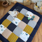 A colorful patchwork quilt with various patterns and textures, including polka dots, stripes, and animal prints, is spread out on a wooden floor. There are also some toys and a basket nearby, suggesting a playful and cozy setting.