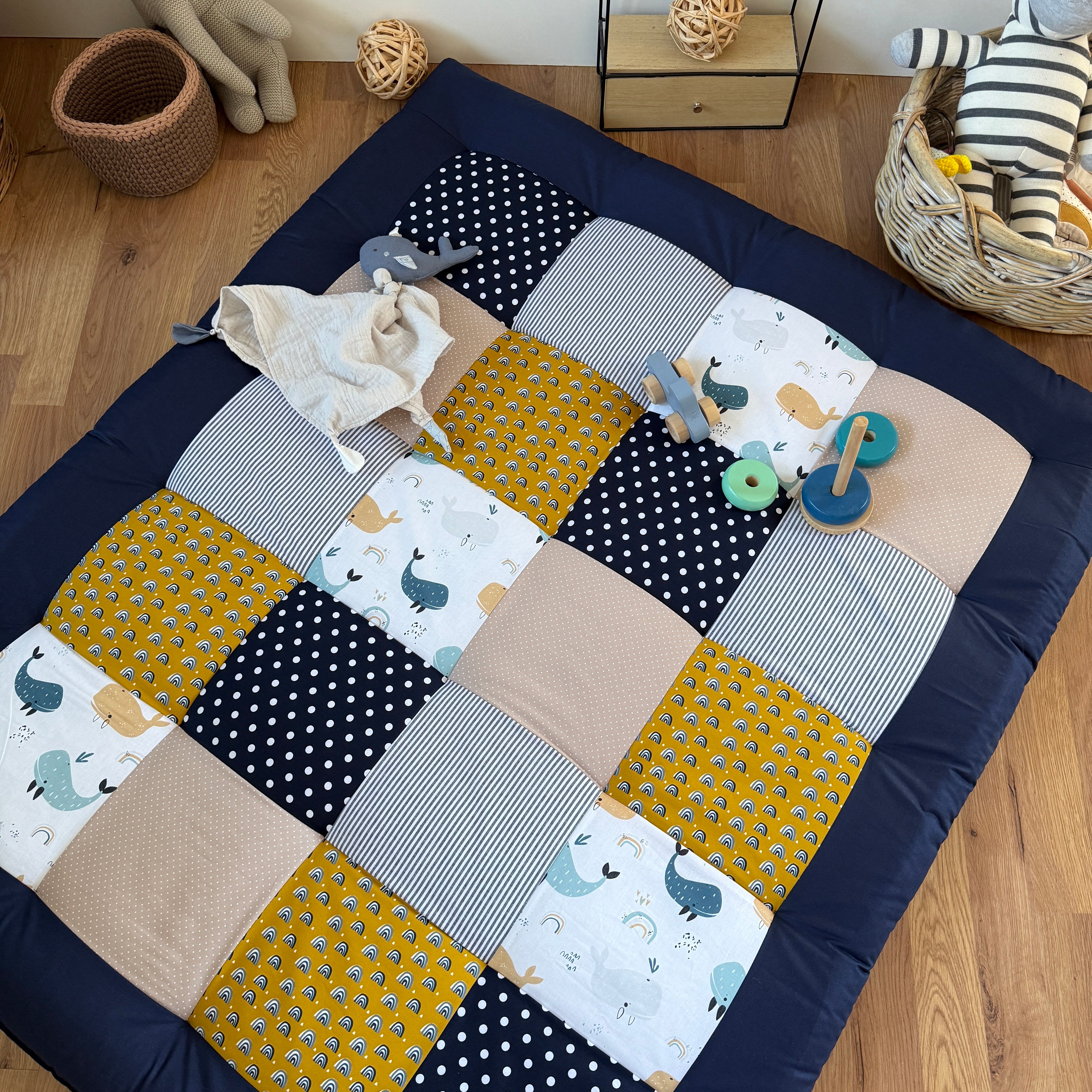 A colorful patchwork quilt with various patterns and textures, including polka dots, stripes, and animal prints, is spread out on a wooden floor. There are also some toys and a basket nearby, suggesting a playful and cozy setting.