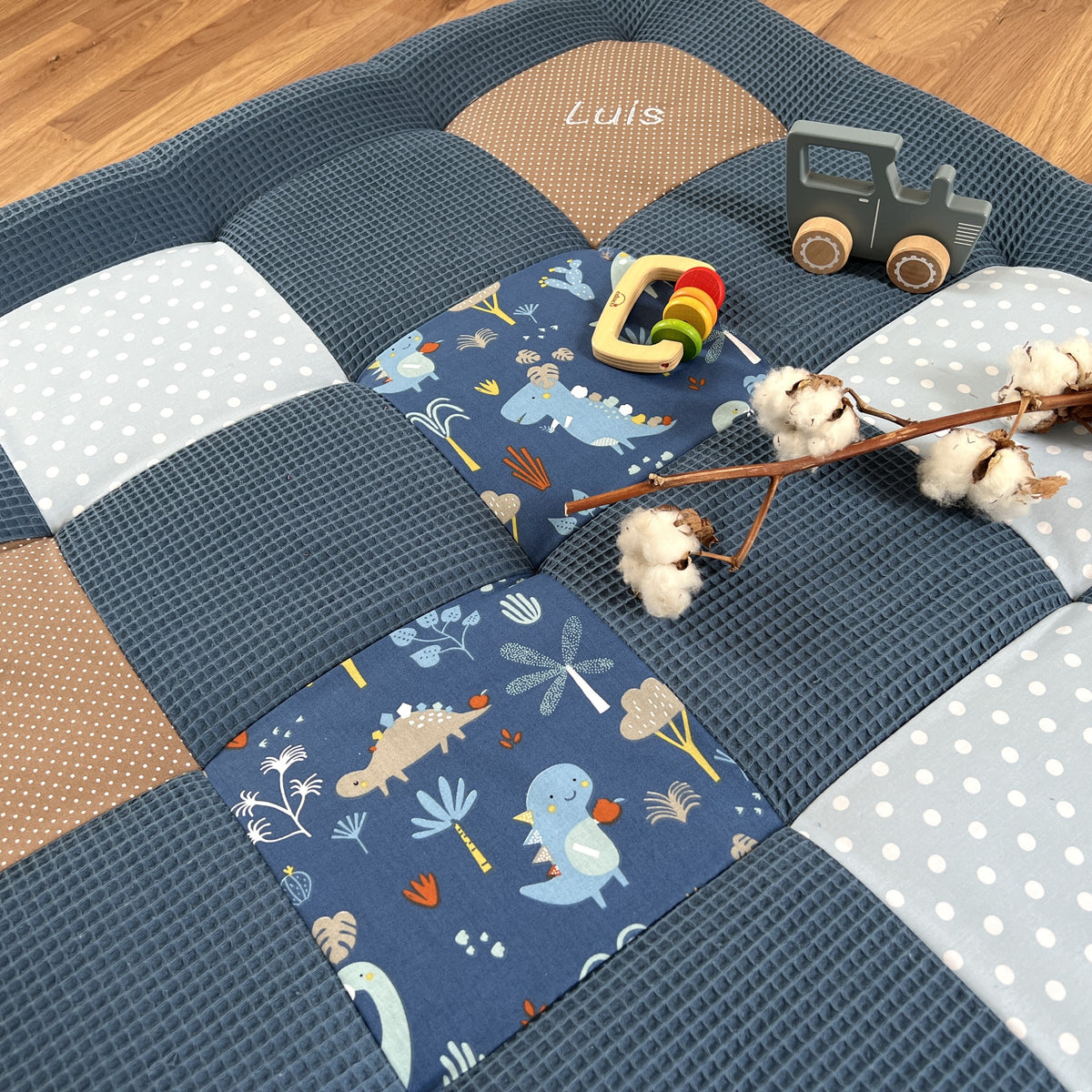 A blue and white checkered baby bedspread with a dinosaur and palm tree pattern, a wooden toy truck, and some cotton balls on the floor.