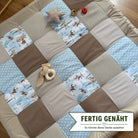A patchwork quilt with various patterns and textures, including polka dots, stripes, and animal prints, is spread out on a wooden floor. There are also a few stuffed animals and a red object placed on the quilt.