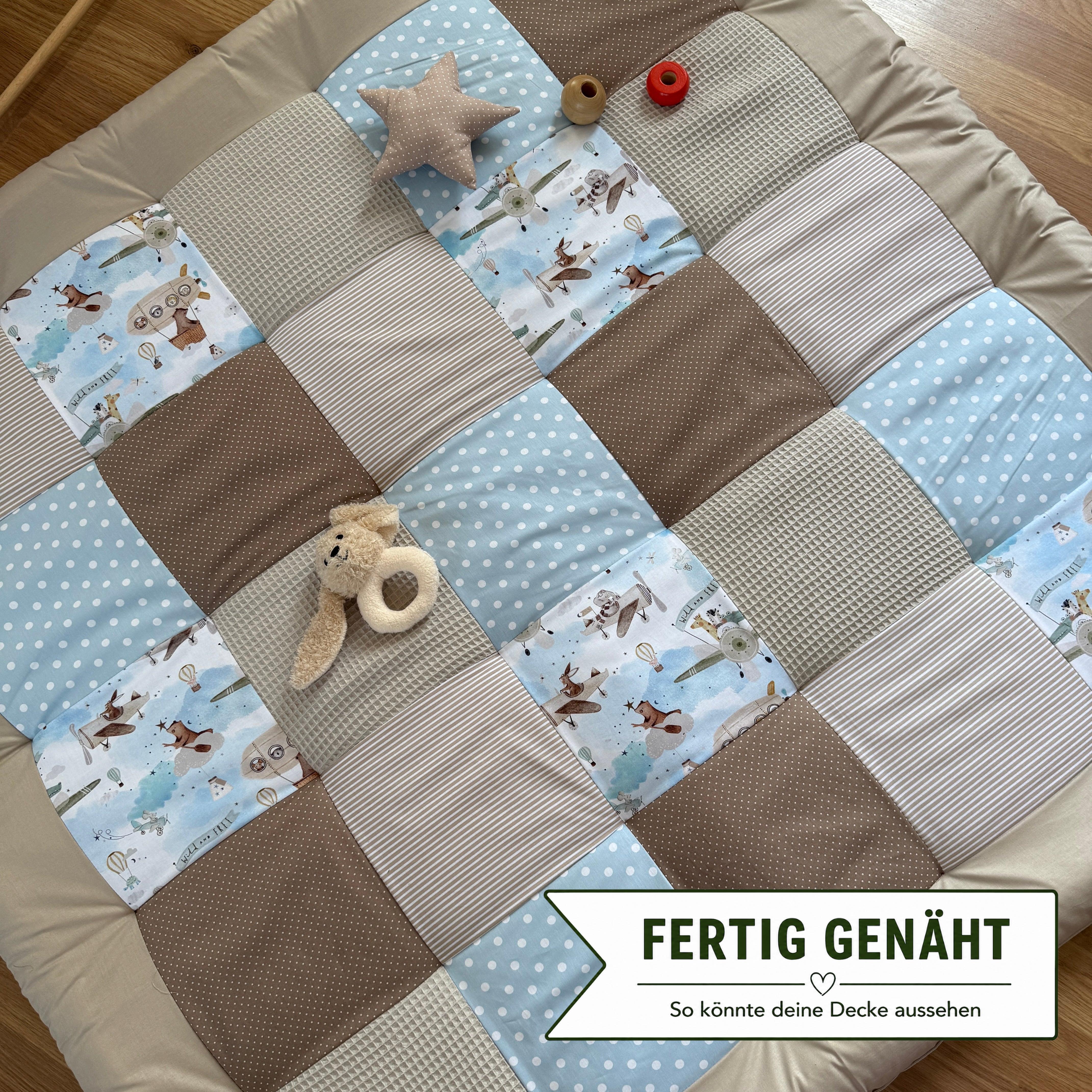 A patchwork quilt with various patterns and textures, including polka dots, stripes, and animal prints, is spread out on a wooden floor. There are also a few stuffed animals and a red object placed on the quilt.