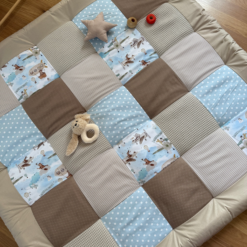 A patchwork quilt with various patterns and textures, including polka dots, stripes, and animal prints, is spread out on a wooden floor. There are also a few stuffed animals and a red object placed on the quilt.