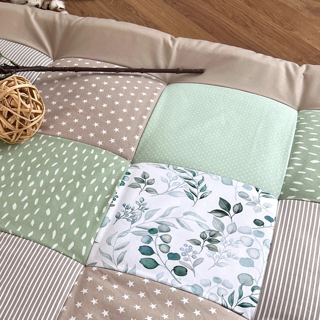 a patchwork quilt with a variety of colors and patterns, including green, white, and brown, laid out on a wooden floor. A small elephant toy is placed next to the quilt, and a wooden crate is also visible in the background.