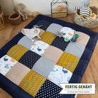 A colorful patchwork quilt with various patterns and textures, including polka dots, stripes, and animal prints, is spread out on a wooden floor. The quilt is surrounded by toys, including a stuffed animal elephant and a toy car. There is also a basket and a wooden toy box in the background.