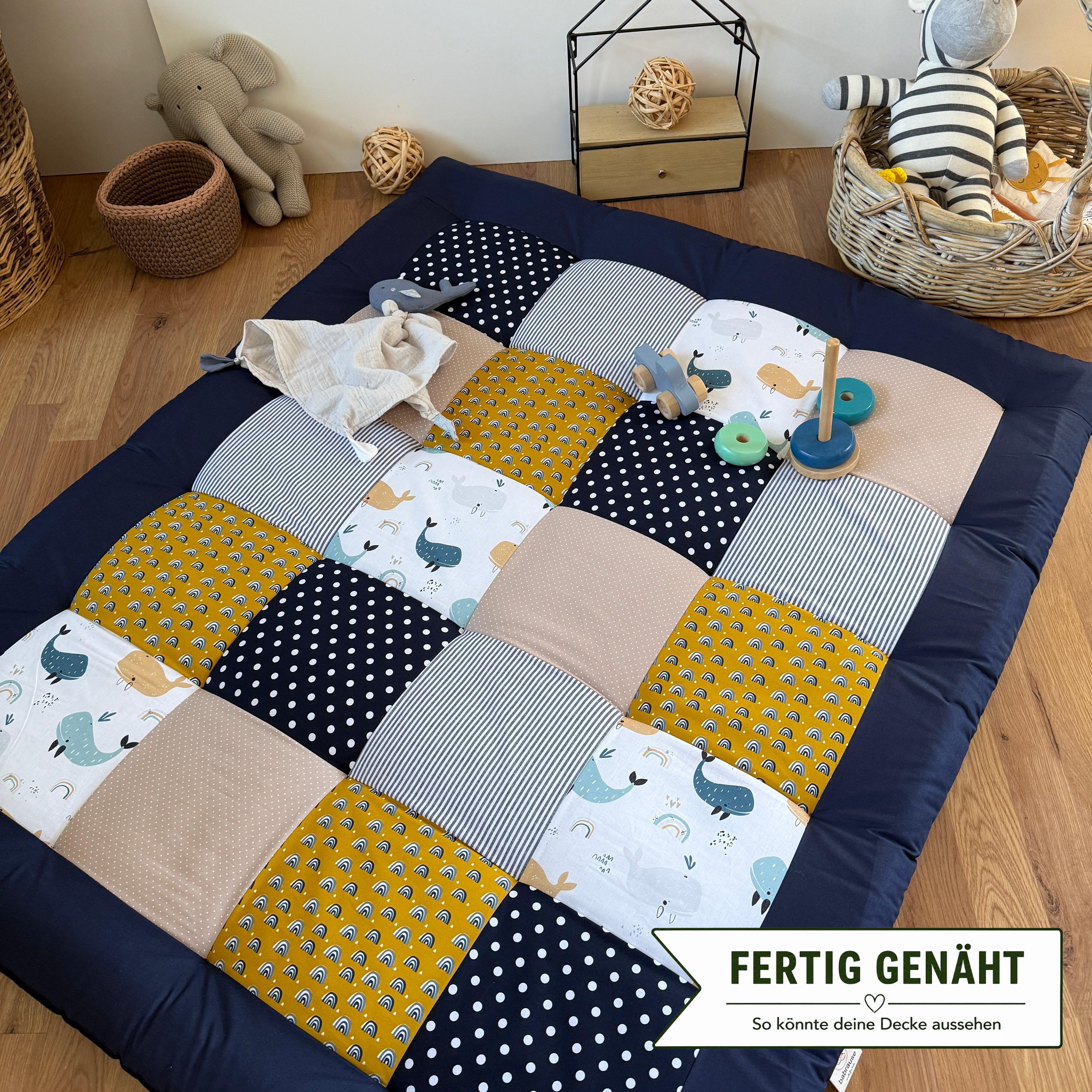 A colorful patchwork quilt with various patterns and textures, including polka dots, stripes, and animal prints, is spread out on a wooden floor. The quilt is surrounded by toys, including a stuffed animal elephant and a toy car. There is also a basket and a wooden toy box in the background.