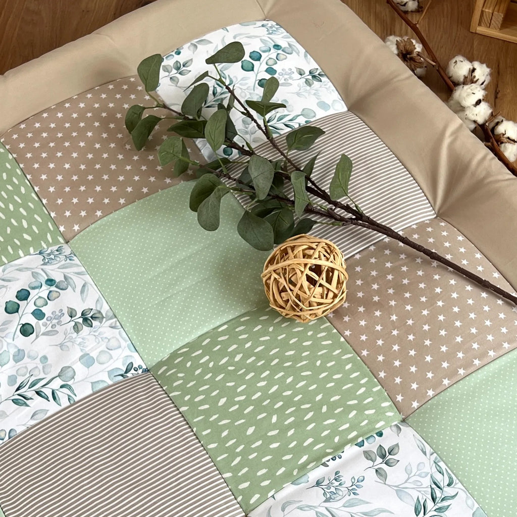 a patchwork quilt with a variety of colors and patterns, including green, brown, and white, laid out on a wooden floor. There is a small stuffed animal, possibly a teddy bear, next to the quilt, and a basket can be seen in the background.