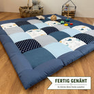 A blue and white quilt-like bedspread with a checkered pattern, placed on a wooden floor. The bedspread has various geometric patterns and polka dots. There are also some toys and a basket in the background.