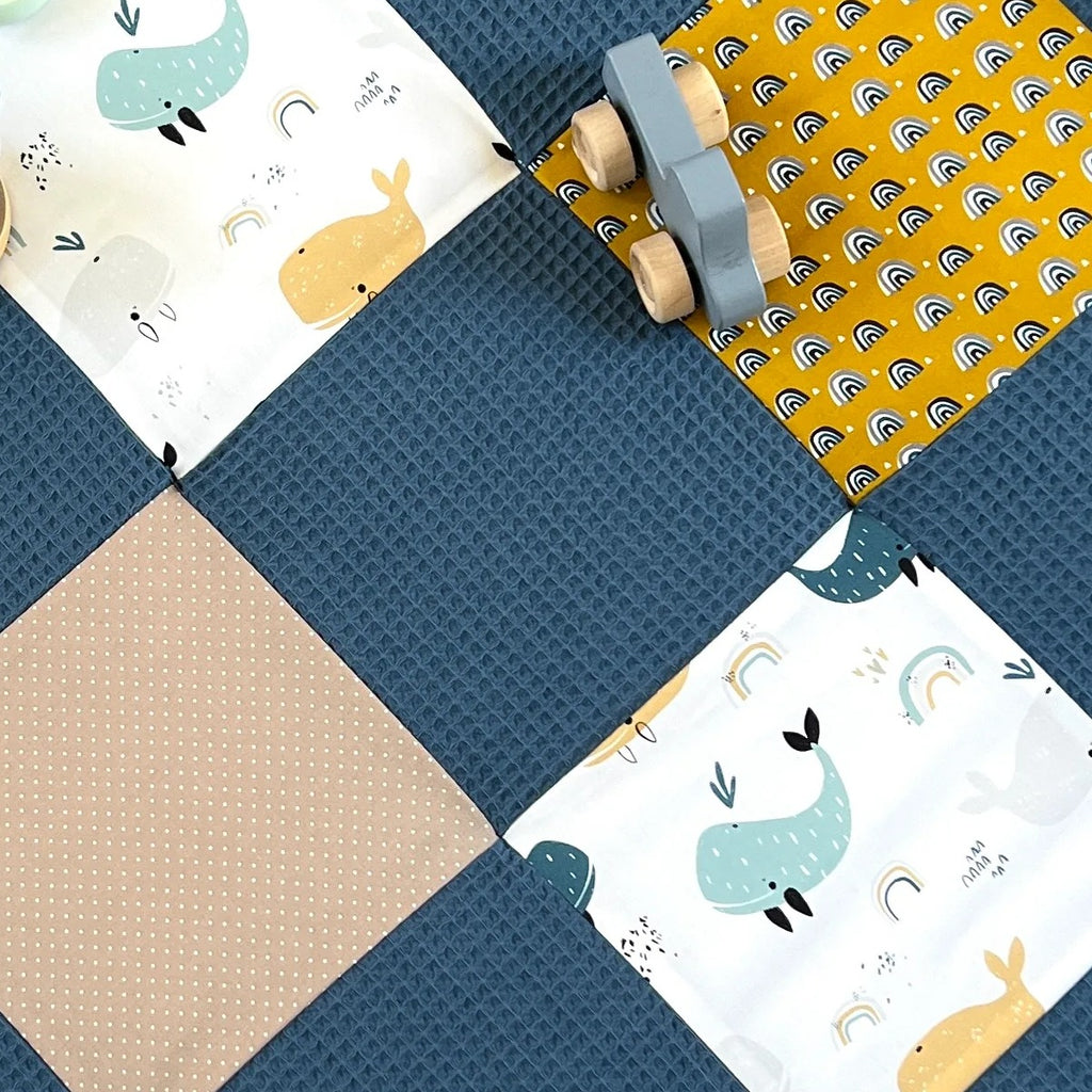 A handmade quilt with a blue and yellow color scheme, featuring a patchwork design with various animal and geometric patterns. The quilt is placed on a wooden floor, and there are some toys and other items scattered around it.
