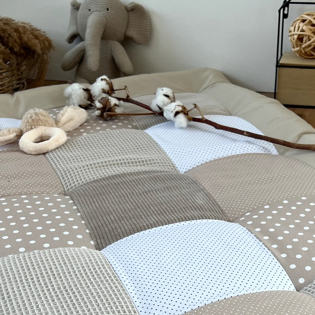 a cozy and inviting space with a patchwork quilt or mat on a wooden floor, featuring a variety of textures and patterns in shades of gray and beige. A small stuffed elephant toy is placed on the mat, and a wooden shelf with a woven basket can be seen in the background.
